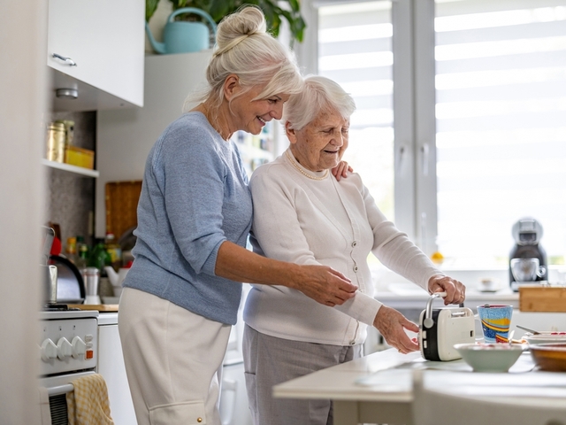 Eine Seniorin und ihre Tochter in mittlerem Alter stehen in der Küche an einem Tisch. Auf dem Tisch stehen Schüsseln vom Frühstück und eine Tasse. Die Seniorin dreht an einem kleinen Radio. Die Tochter steht hinter ihr, hat einen Arm auf ihre Schulter gelegt. Beide lächeln. 