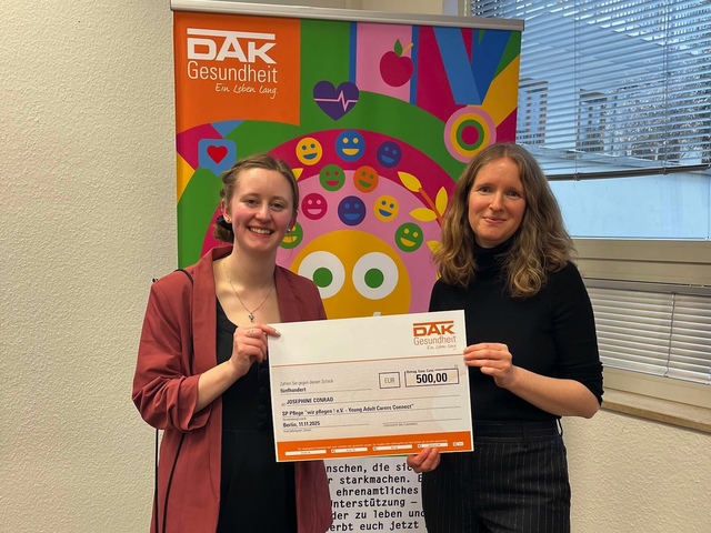 Josephine Conrad und Lisa Thelen stehen vor einem Banner des DAK Wettbewerbs "Gesichter für ein gesundes Miteinander". Sie halten den Check über das Preisgeld in den Händen. Sie lächeln in die Kamera.